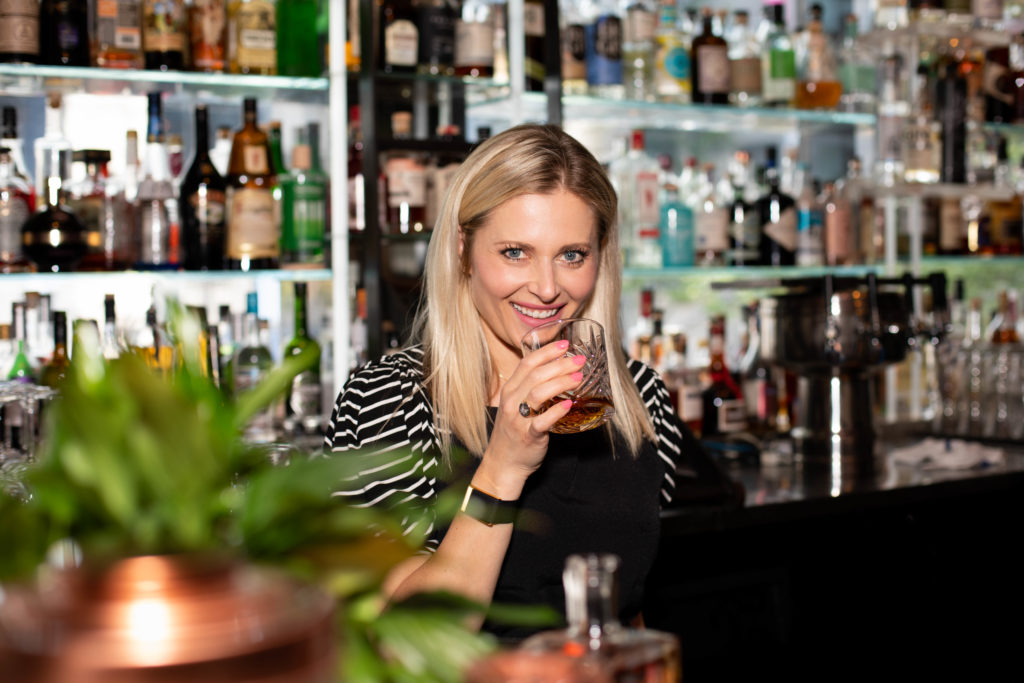 Portrait of Heritage 1904 Owner and Operator, Missy Firnstahl-Claridge. A blonde woman is standing in front of a well-stocked bar holding an etched glass partially filled with Bourbon in her right hand. She is smiling and looking pleasant.
